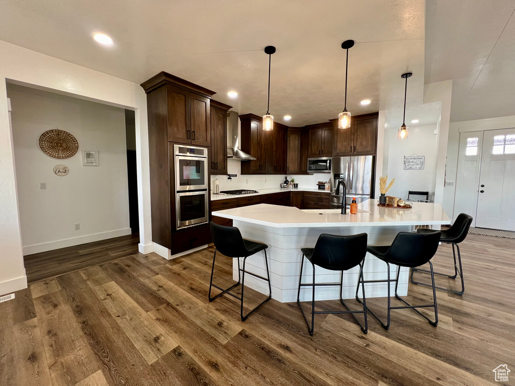 Kitchen with dark wood-type flooring, a large island with sink, stainless steel appliances, and decorative light fixtures