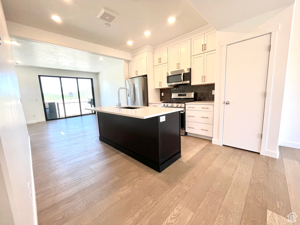 Kitchen with stainless steel appliances, light wood-style flooring, a center island with sink, white cabinets, and tasteful backsplash