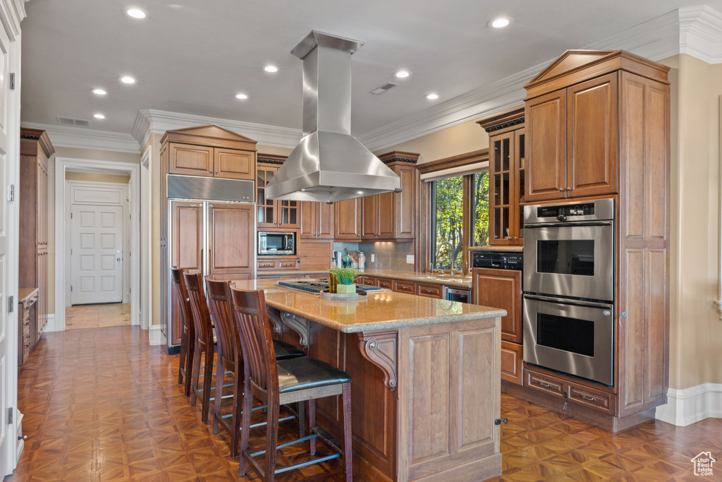 Kitchen with light stone counters, built in appliances, an island with sink, island exhaust hood, and crown molding