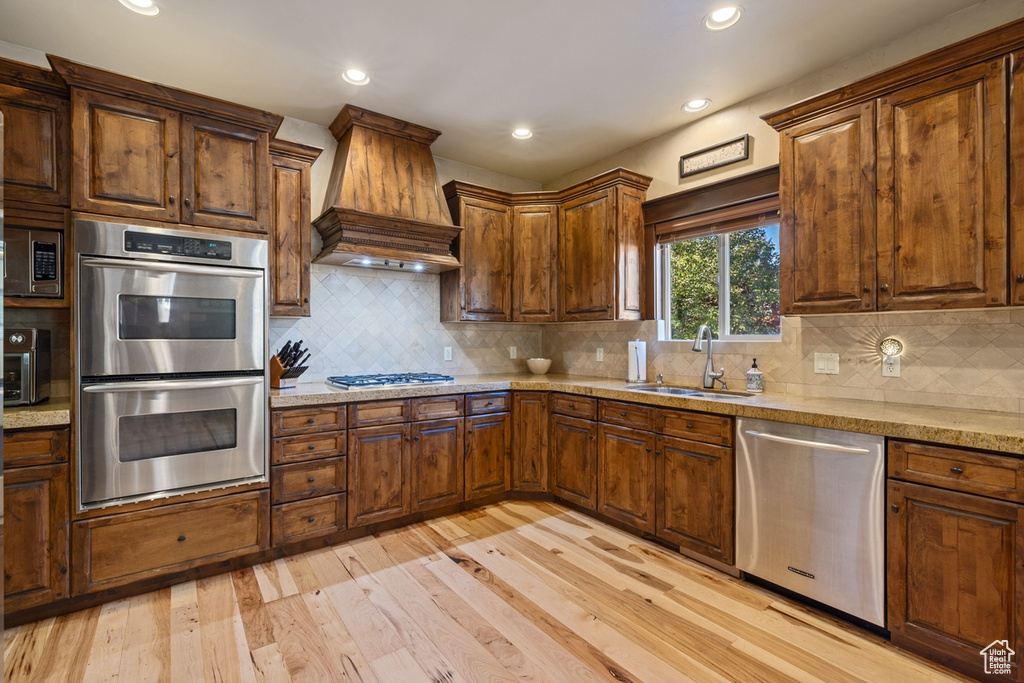 Kitchen featuring stainless steel appliances, decorative backsplash, sink, custom exhaust hood, and light hardwood / wood-style flooring