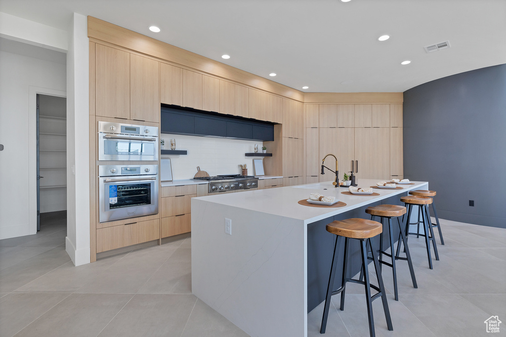 Kitchen with a kitchen bar, a kitchen island with sink, light tile patterned flooring, light brown cabinets, and appliances with stainless steel finishes