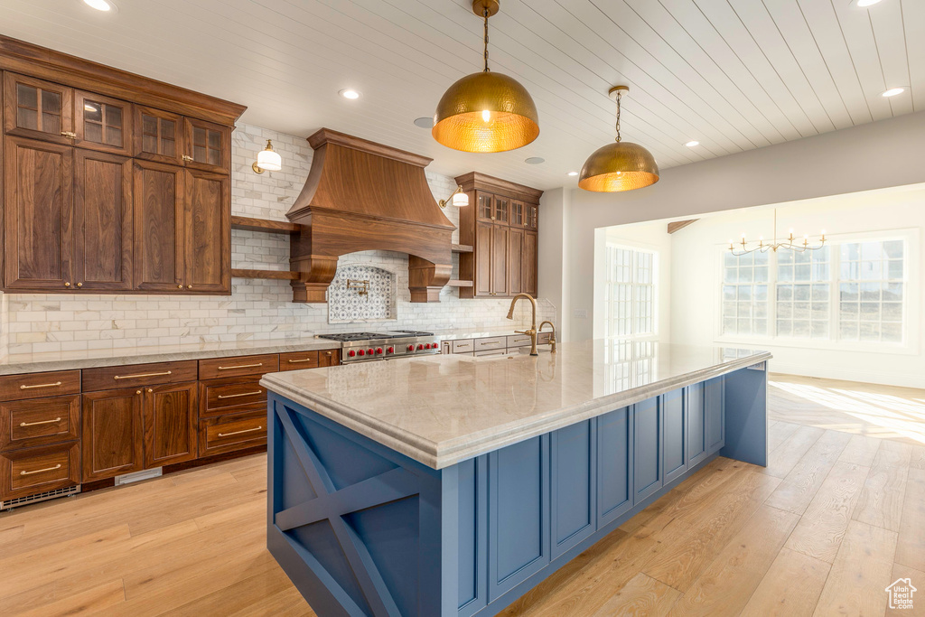 Kitchen featuring light hardwood / wood-style floors, sink, premium range hood, a kitchen island with sink, and pendant lighting