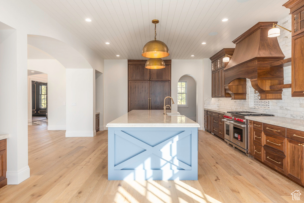 Kitchen with range with two ovens, hanging light fixtures, tasteful backsplash, an island with sink, and light wood-type flooring