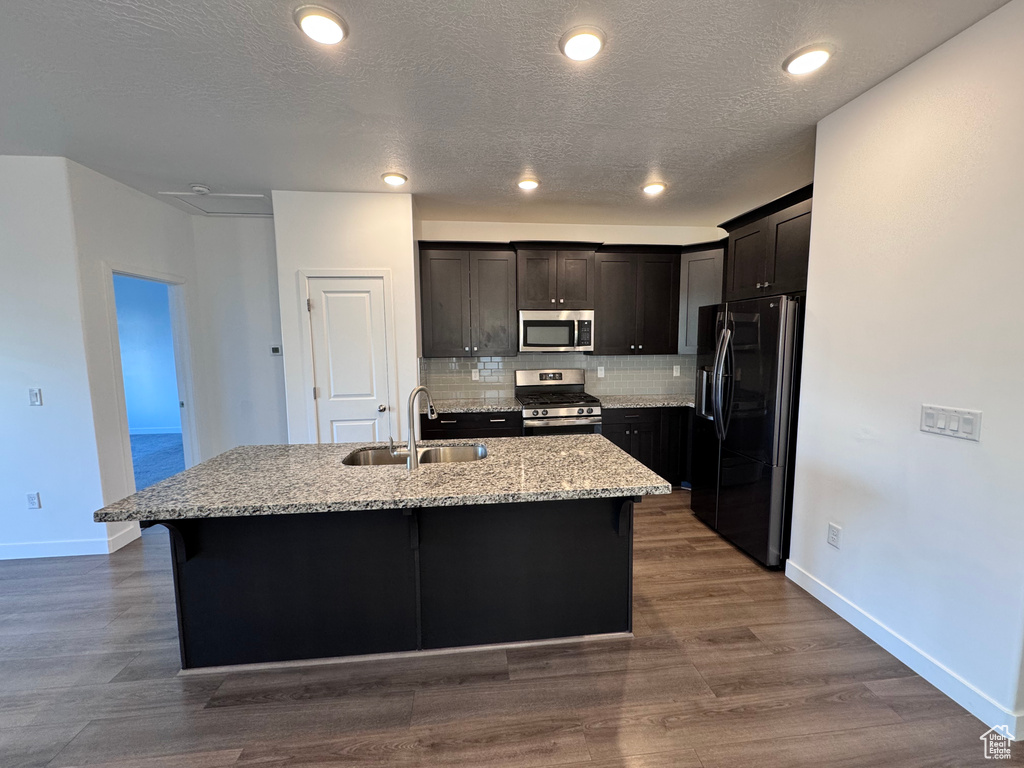 Kitchen featuring an island with sink, a sink, tasteful backsplash, stainless steel appliances, and dark wood-type flooring