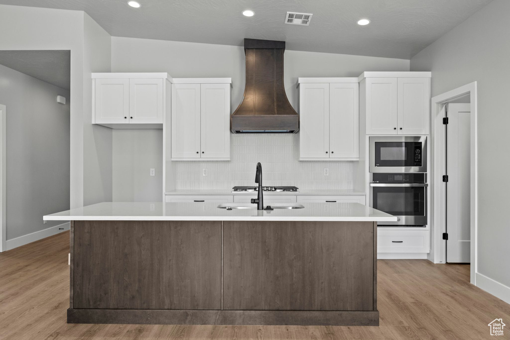Kitchen featuring stainless steel appliances, a kitchen island with sink, white cabinets, and custom range hood