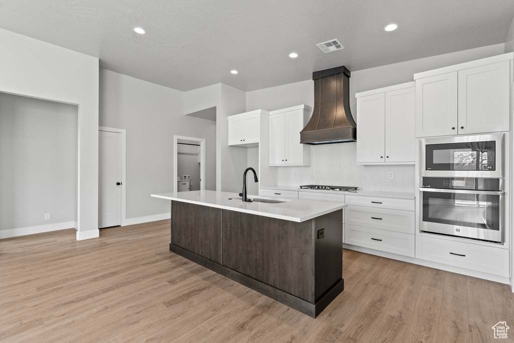 Kitchen with a sink, light countertops, an island with sink, custom range hood, and stovetop