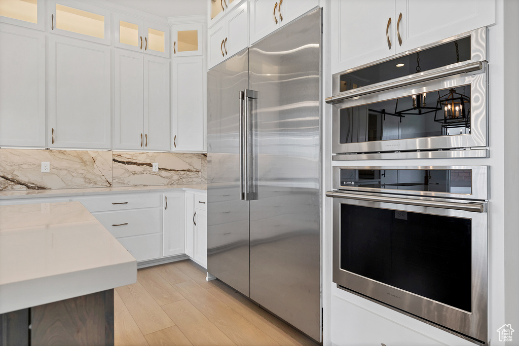 Kitchen with stainless steel appliances, white cabinets, light wood-style floors, glass insert cabinets, and tasteful backsplash