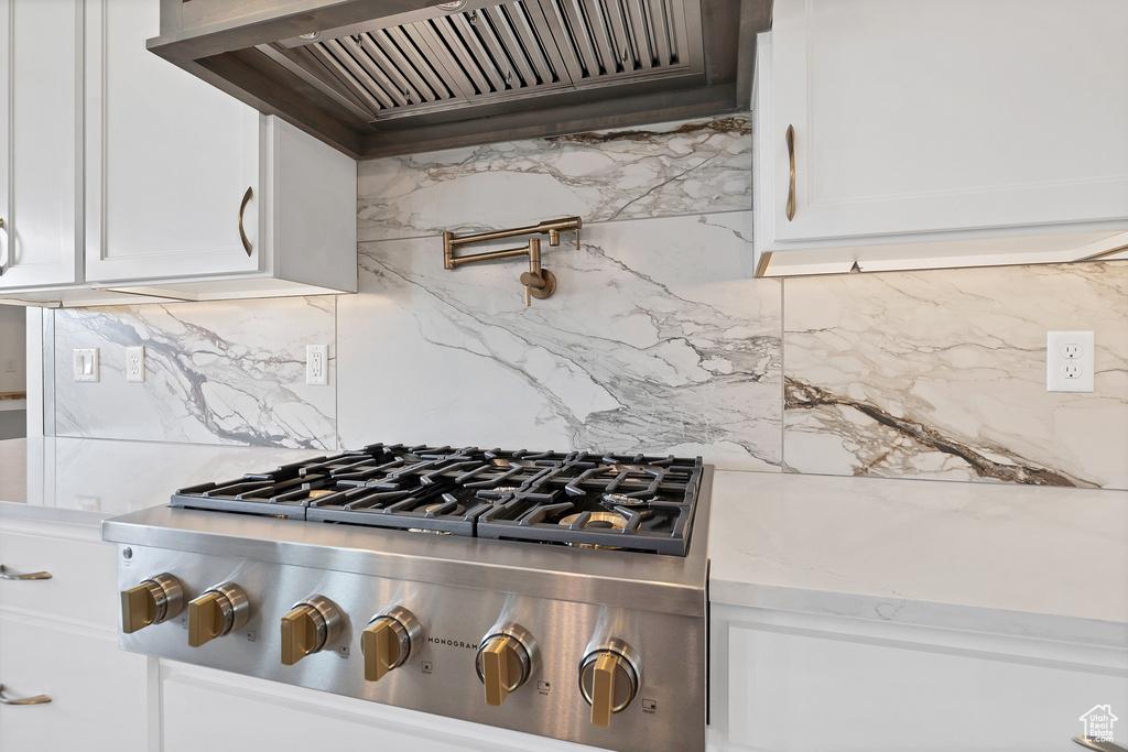 Kitchen view of stainless steel gas cooktop, custom range hood, pot filler, decorative backsplash, and white cabinets