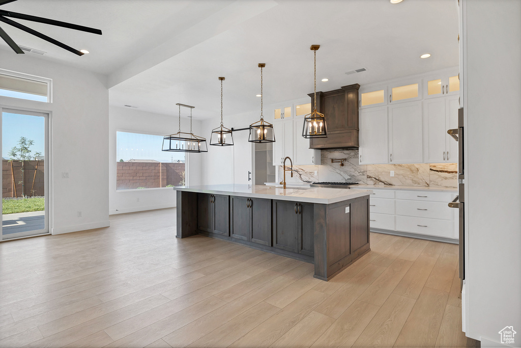 Kitchen featuring light countertops, decorative backsplash, a kitchen island with sink, white cabinets, and light wood finished floors