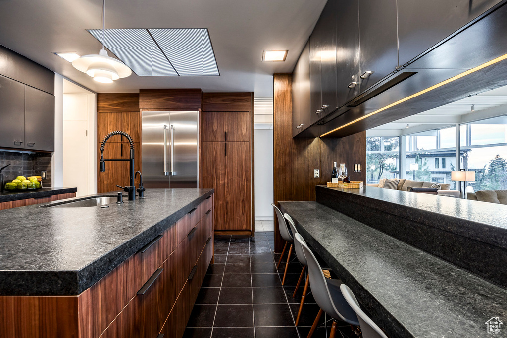 Kitchen with backsplash, stainless steel built in refrigerator, a kitchen island with sink, sink, and dark tile patterned flooring