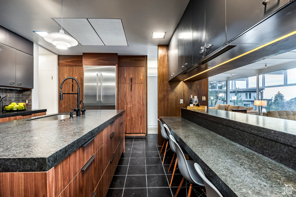 Kitchen with sink, backsplash, stainless steel built in fridge, dark tile patterned flooring, and a center island with sink