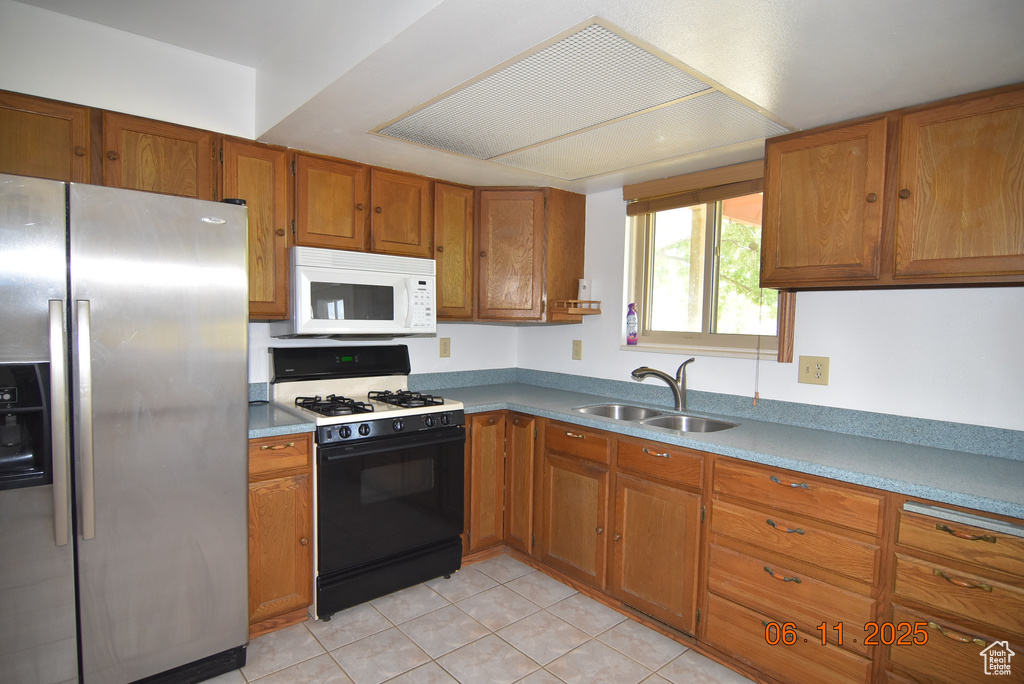 Kitchen with stainless steel fridge, range with gas stovetop, white microwave, brown cabinetry, and light tile patterned flooring