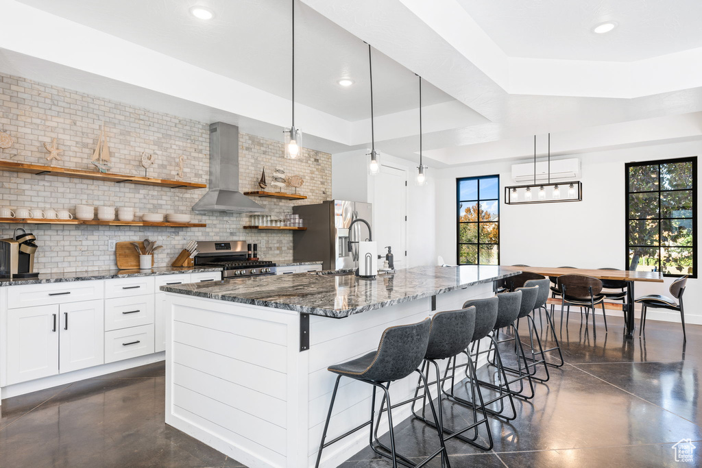 Kitchen featuring wall chimney exhaust hood, stainless steel appliances, tasteful backsplash, decorative light fixtures, and a tray ceiling