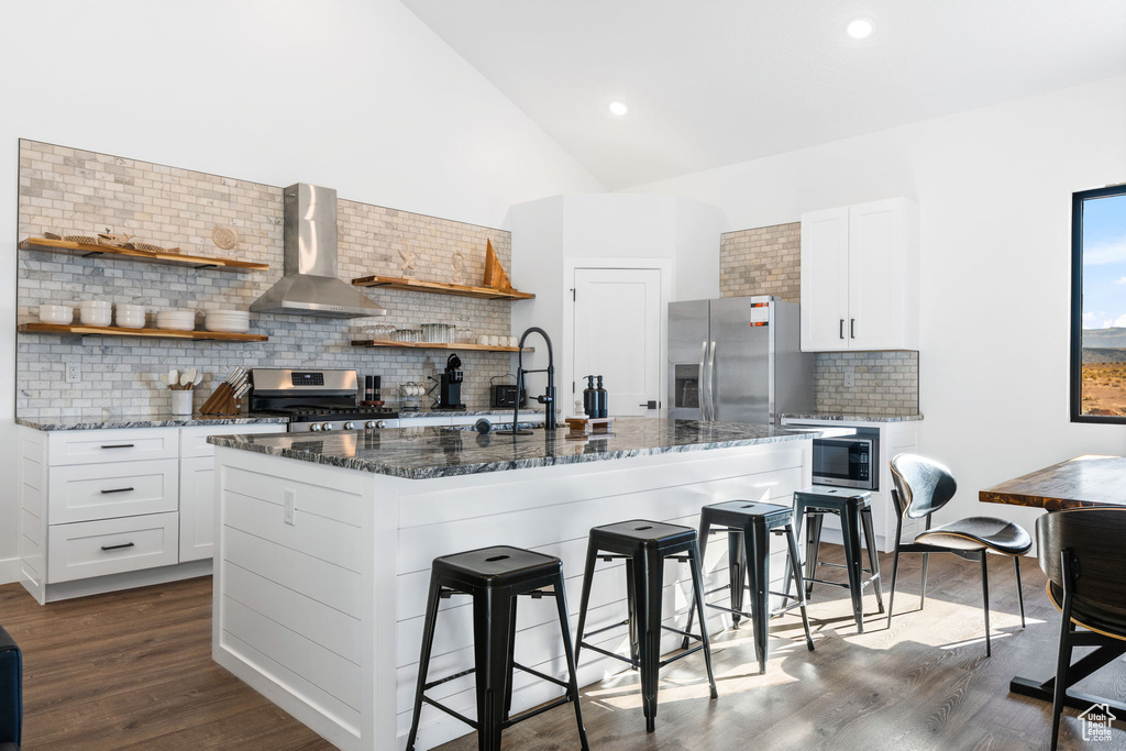 Kitchen featuring a center island with sink, wall chimney exhaust hood, white cabinetry, and appliances with stainless steel finishes