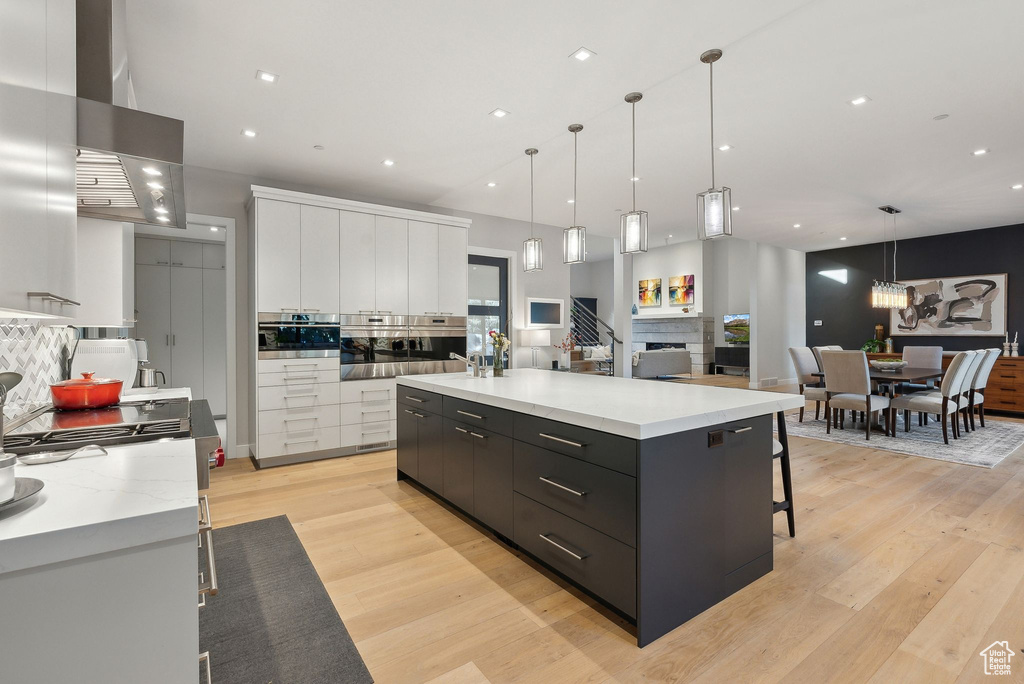 Kitchen with modern cabinets, wall chimney exhaust hood, white cabinets, light wood-style floors, and a large island with sink