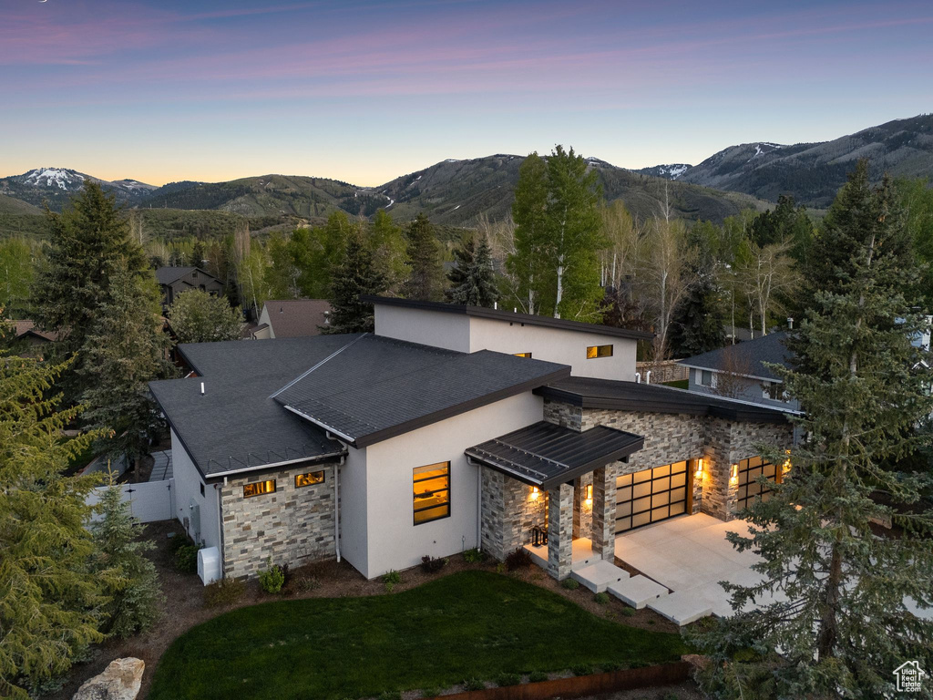 Back of house at dusk with stone siding, a mountain view, driveway, and stucco siding