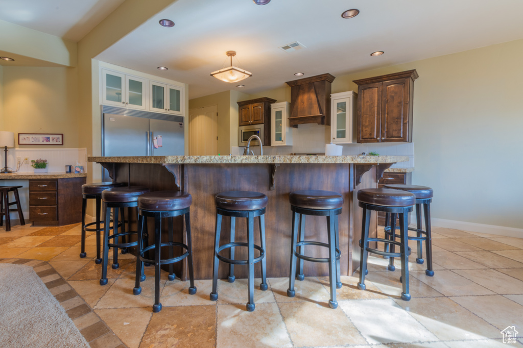 Kitchen featuring built in appliances, an island with sink, a breakfast bar area, dark brown cabinets, and custom range hood