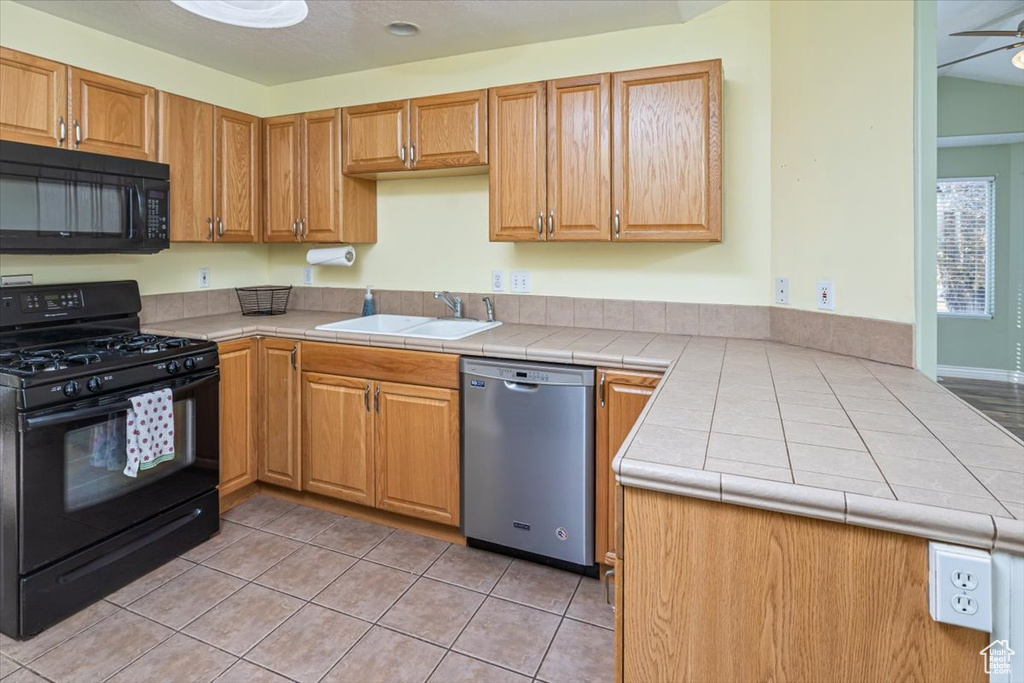 Kitchen with tile countertops, black appliances, sink, ceiling fan, and light tile patterned flooring