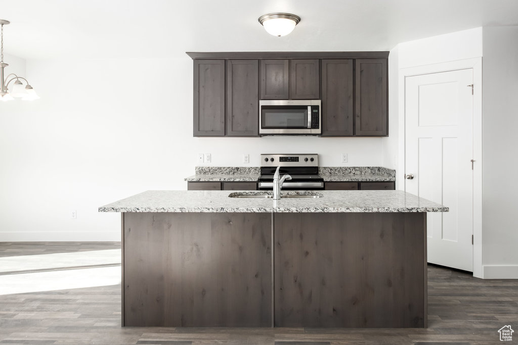 Kitchen featuring dark wood-type flooring, an island with sink, light stone counters, dark brown cabinetry, and stainless steel appliances