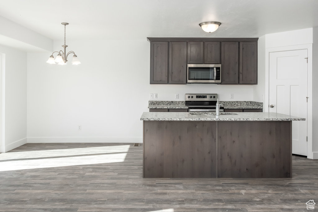 Kitchen featuring dark brown cabinetry, dark wood-type flooring, hanging light fixtures, light stone counters, and appliances with stainless steel finishes