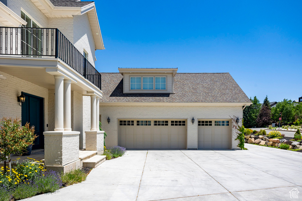 View of property exterior featuring driveway, brick siding, a balcony, roof with shingles, and an attached garage