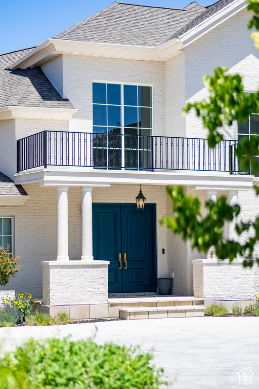 Doorway to property featuring a balcony, brick siding, covered porch, and roof with shingles