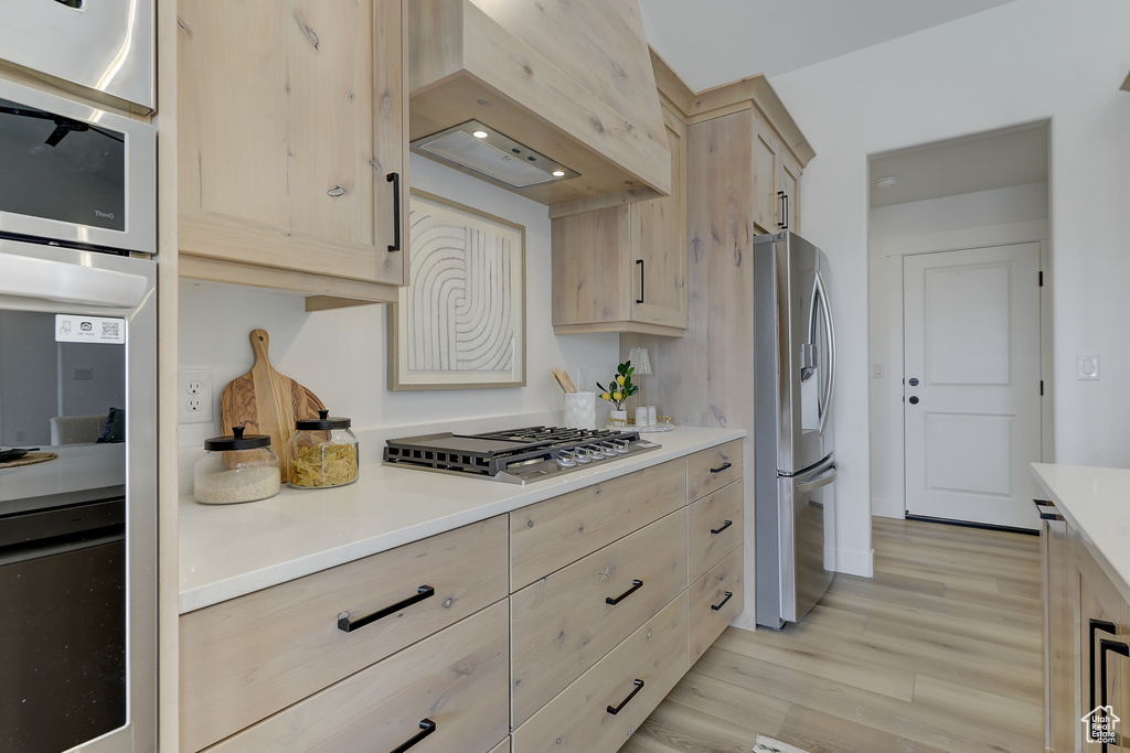 Kitchen with light brown cabinetry, custom exhaust hood, stainless steel appliances, and light wood-type flooring