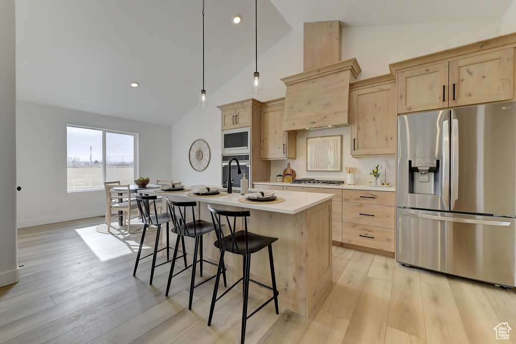 Kitchen with light brown cabinetry, decorative light fixtures, appliances with stainless steel finishes, a kitchen island with sink, and light hardwood / wood-style floors