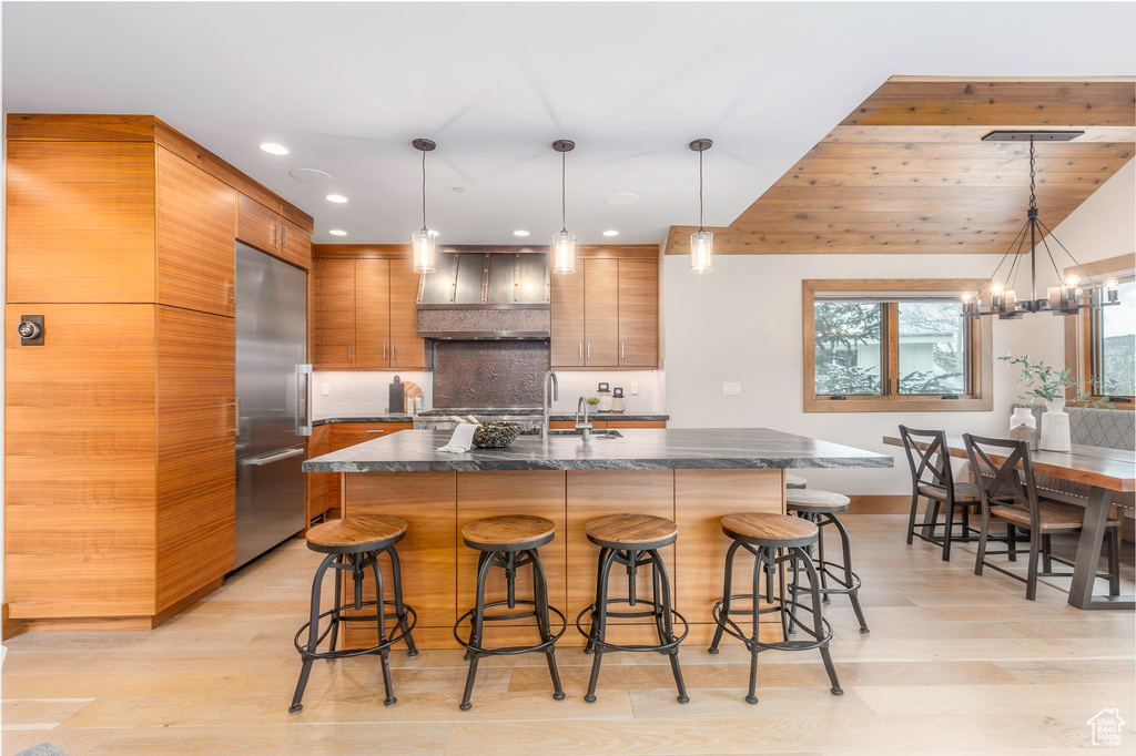 Kitchen featuring stainless steel built in fridge, range hood, dark countertops, light wood-type flooring, and a breakfast bar