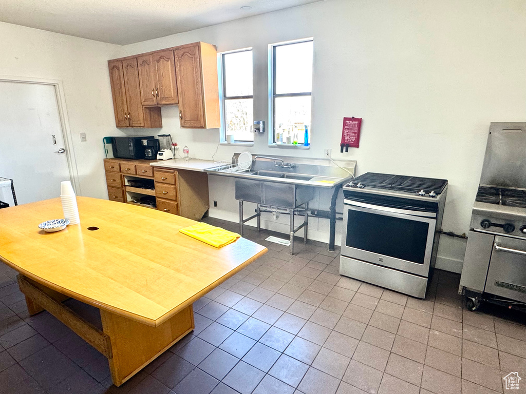 Kitchen with stainless steel gas stove, light tile patterned flooring, black microwave, light countertops, and brown cabinets