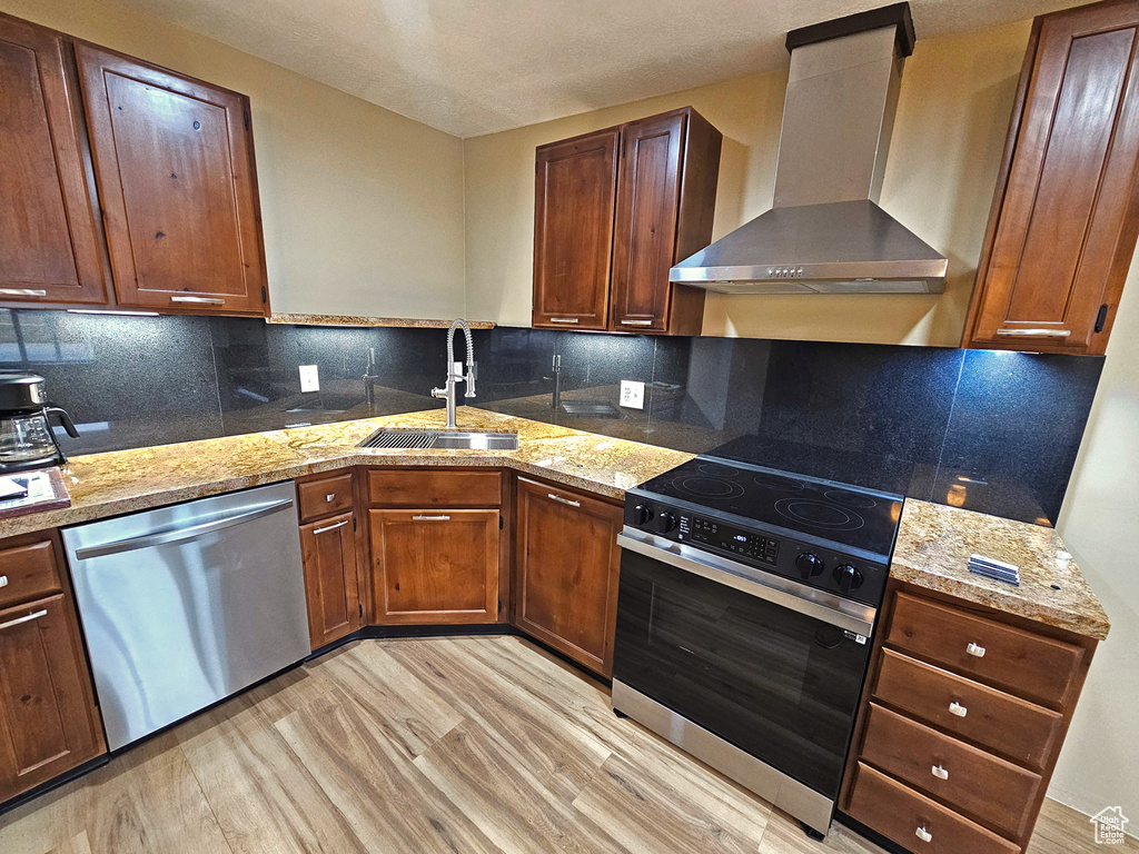 Kitchen featuring wall chimney range hood, sink, appliances with stainless steel finishes, decorative backsplash, and light wood-type flooring