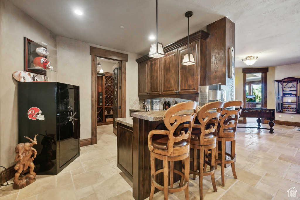 Kitchen with stone tile flooring, dark brown cabinets, a breakfast bar, decorative light fixtures, and light countertops