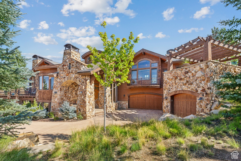 View of front facade featuring stone siding, a balcony, a garage, concrete driveway, and a pergola