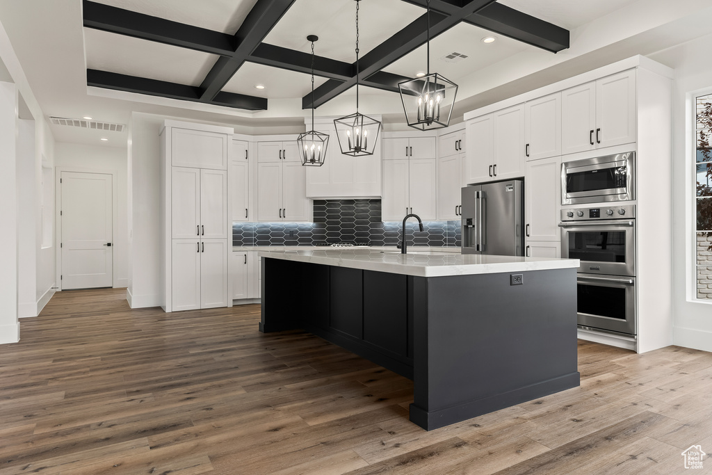 Kitchen with backsplash, appliances with stainless steel finishes, an island with sink, white cabinets, and hardwood / wood-style flooring