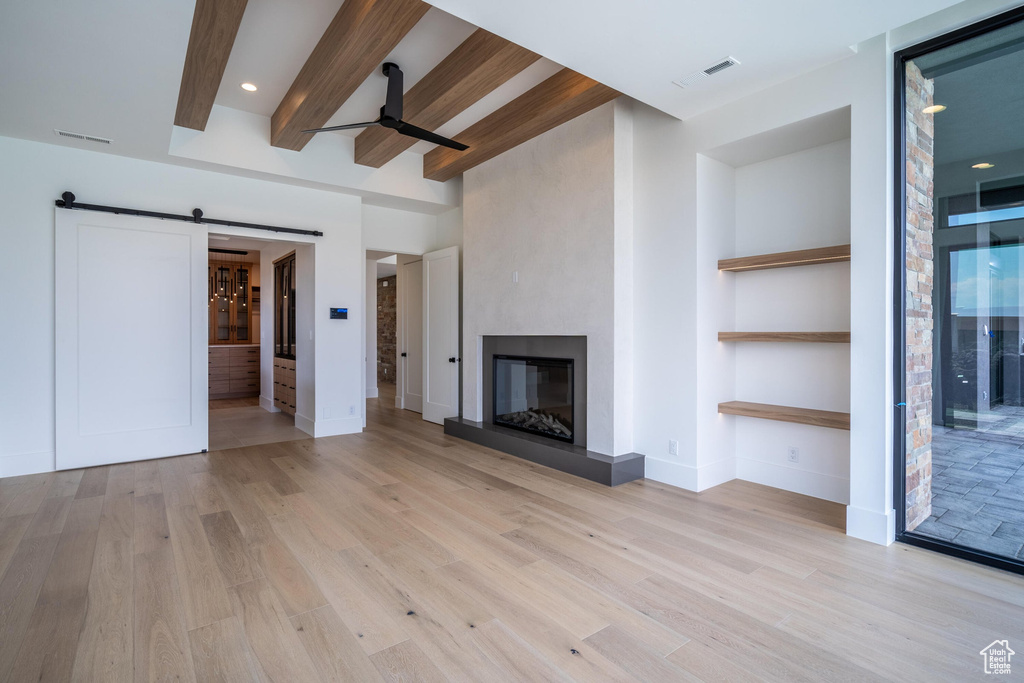 Unfurnished living room with ceiling fan, a barn door, light hardwood / wood-style floors, beam ceiling, and a large fireplace