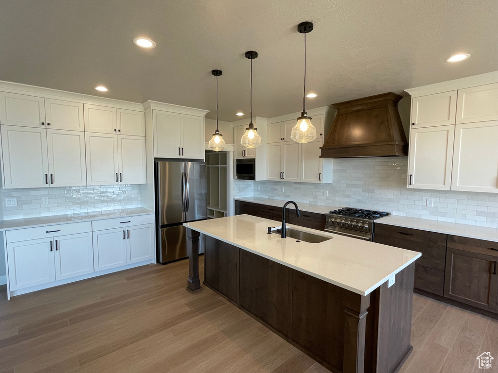 Kitchen featuring custom exhaust hood, stainless steel appliances, a kitchen island with sink, sink, and pendant lighting