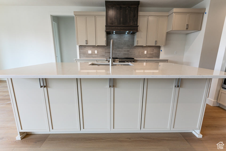 Kitchen with an island with sink, premium range hood, a sink, light wood-style flooring, and backsplash