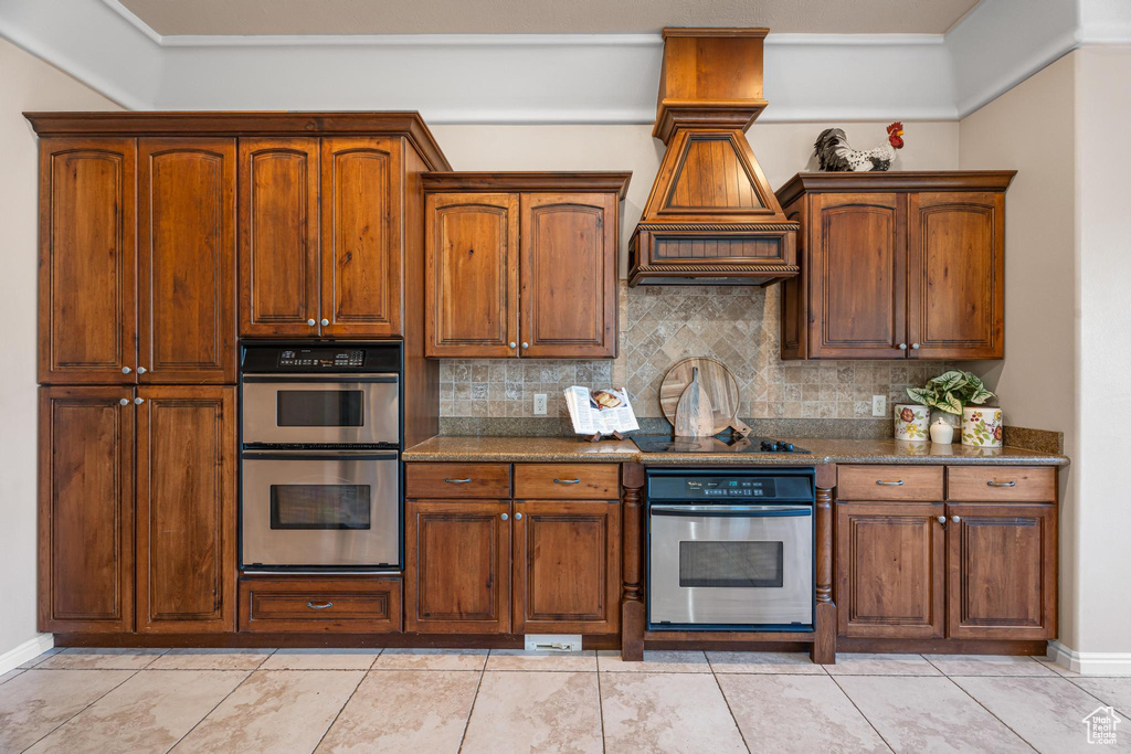 Kitchen with double oven, dark stone countertops, black electric cooktop, decorative backsplash, and light tile patterned floors