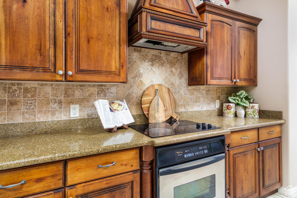 Kitchen with premium range hood, black electric stovetop, oven, decorative backsplash, and light stone counters