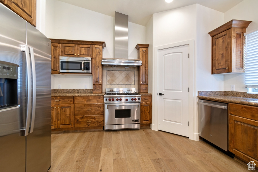 Kitchen with appliances with stainless steel finishes, tasteful backsplash, wall chimney range hood, light hardwood / wood-style flooring, and stone counters