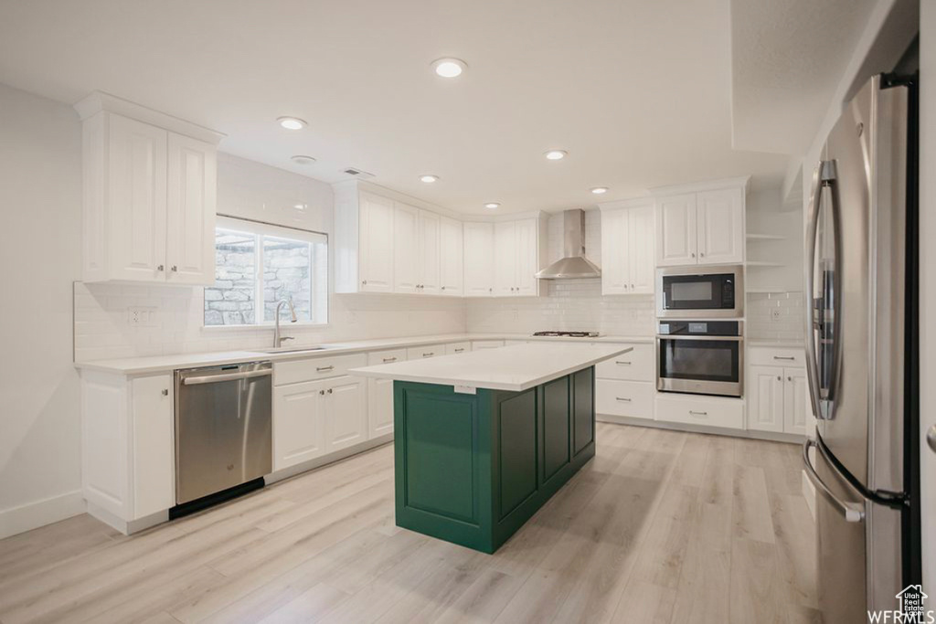 Kitchen with wall chimney exhaust hood, a kitchen island, appliances with stainless steel finishes, white cabinets, and light wood-type flooring