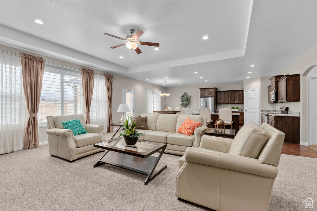 Living room with a tray ceiling, light colored carpet, and ceiling fan with notable chandelier