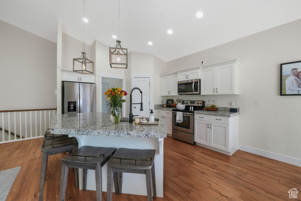 Kitchen featuring appliances with stainless steel finishes, light stone counters, a center island with sink, decorative light fixtures, and white cabinets