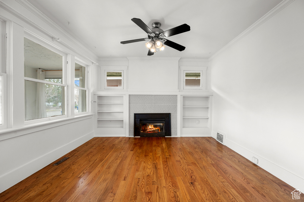 Unfurnished living room with light wood-type flooring, ornamental molding, built in shelves, ceiling fan, and a tiled fireplace