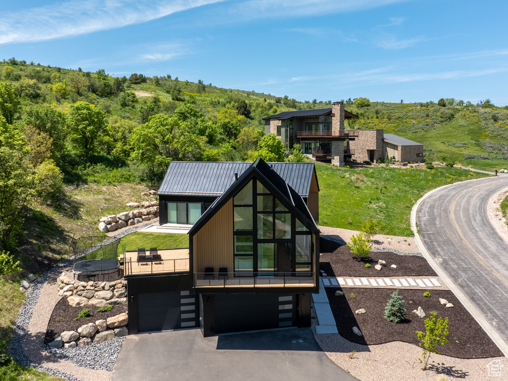 Back of property featuring asphalt driveway, an attached garage, metal roof, a view of trees, and a balcony