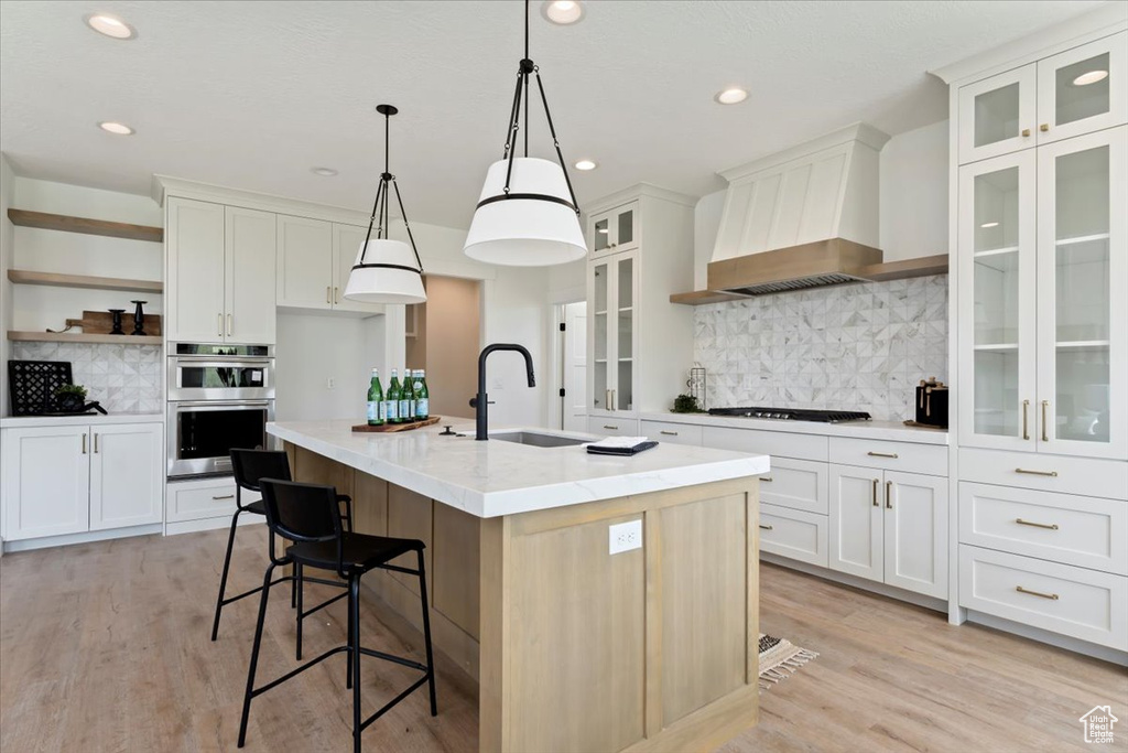 Kitchen featuring decorative backsplash, an island with sink, double oven, decorative light fixtures, and custom range hood