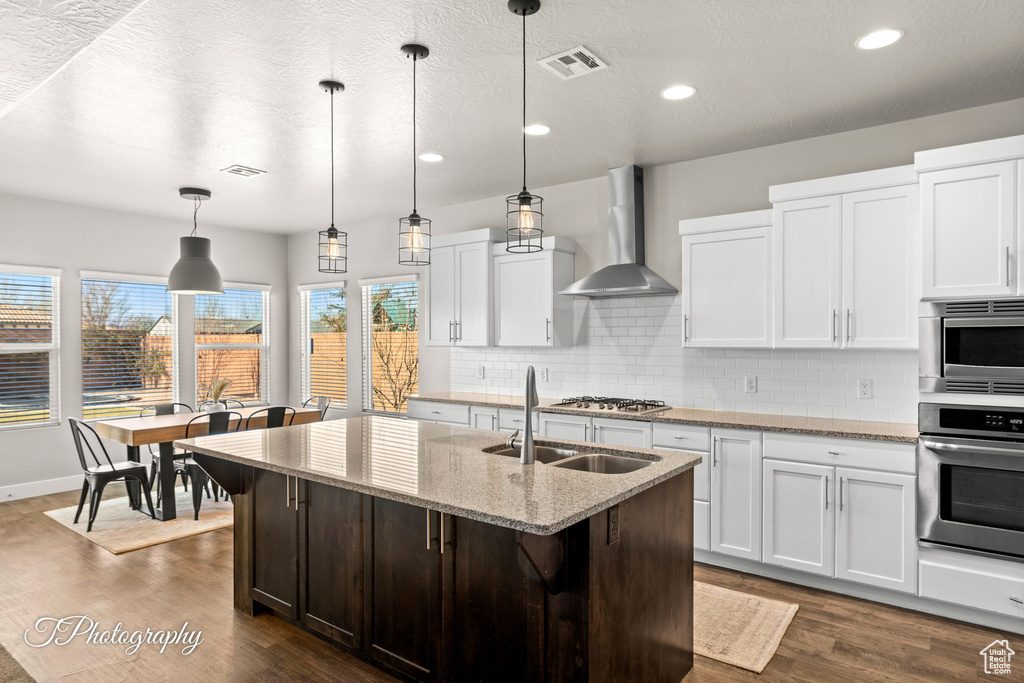 Kitchen featuring light stone countertops, stainless steel appliances, wall chimney range hood, a kitchen island with sink, and white cabinets