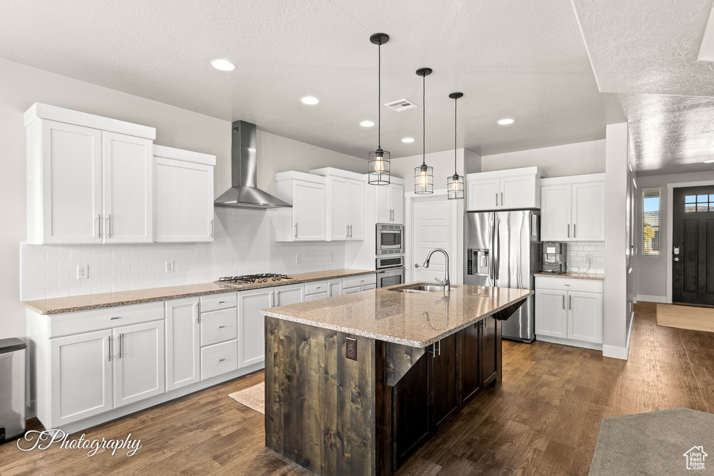 Kitchen featuring a kitchen island with sink, white cabinets, sink, wall chimney exhaust hood, and appliances with stainless steel finishes