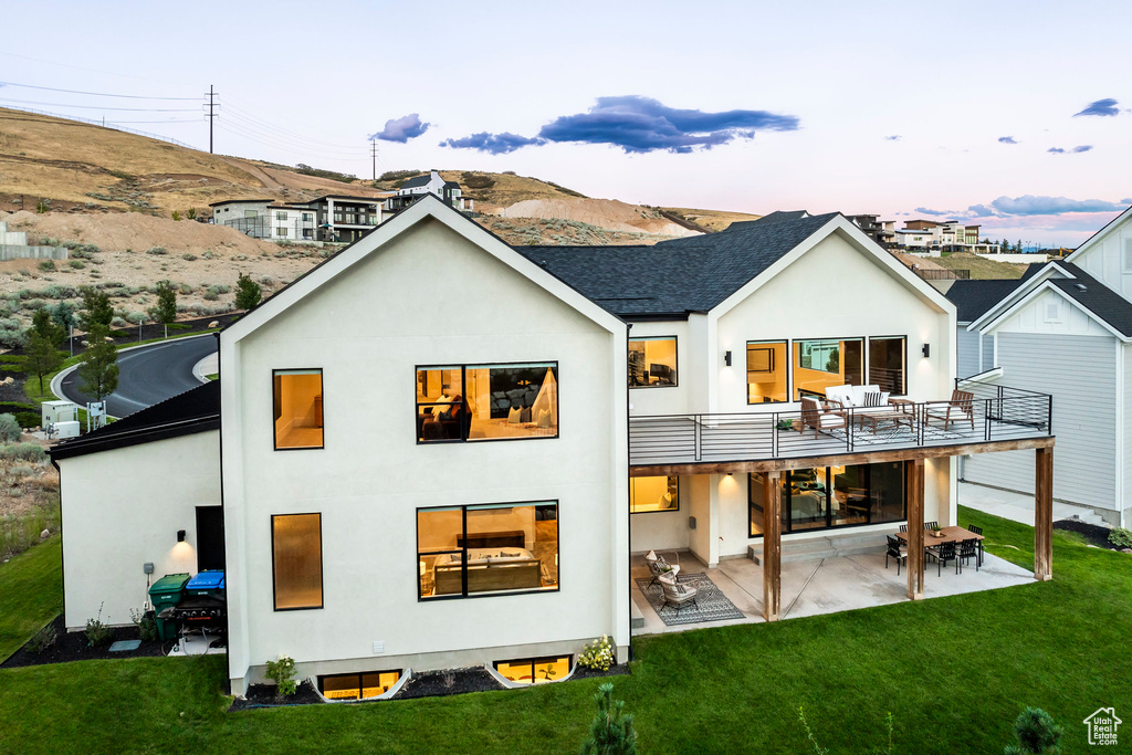 Back house at dusk with a mountain view, a balcony, a patio area, and a lawn
