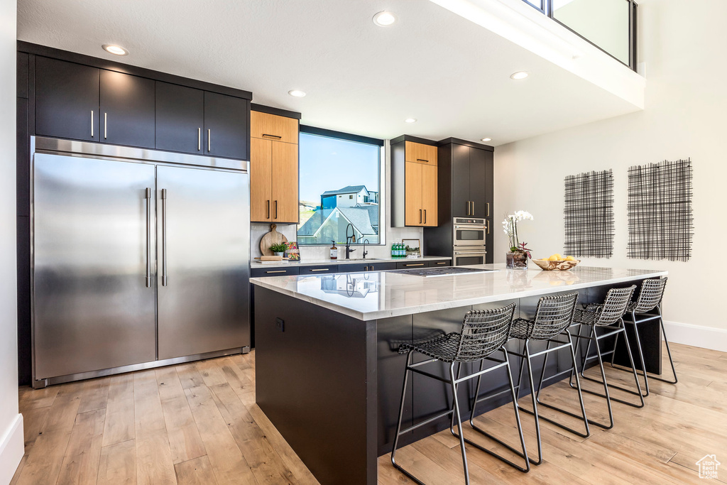 Kitchen with a kitchen bar, light stone countertops, stainless steel built in refrigerator, light hardwood / wood-style flooring, and a center island